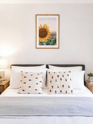 Ein ordentlich bezogenes Bett mit weißer Bettwäsche und gemusterten Kissen, ein gerahmtes Bild mit Sonnenblumen an der Wand und eine Nachttischlampe.