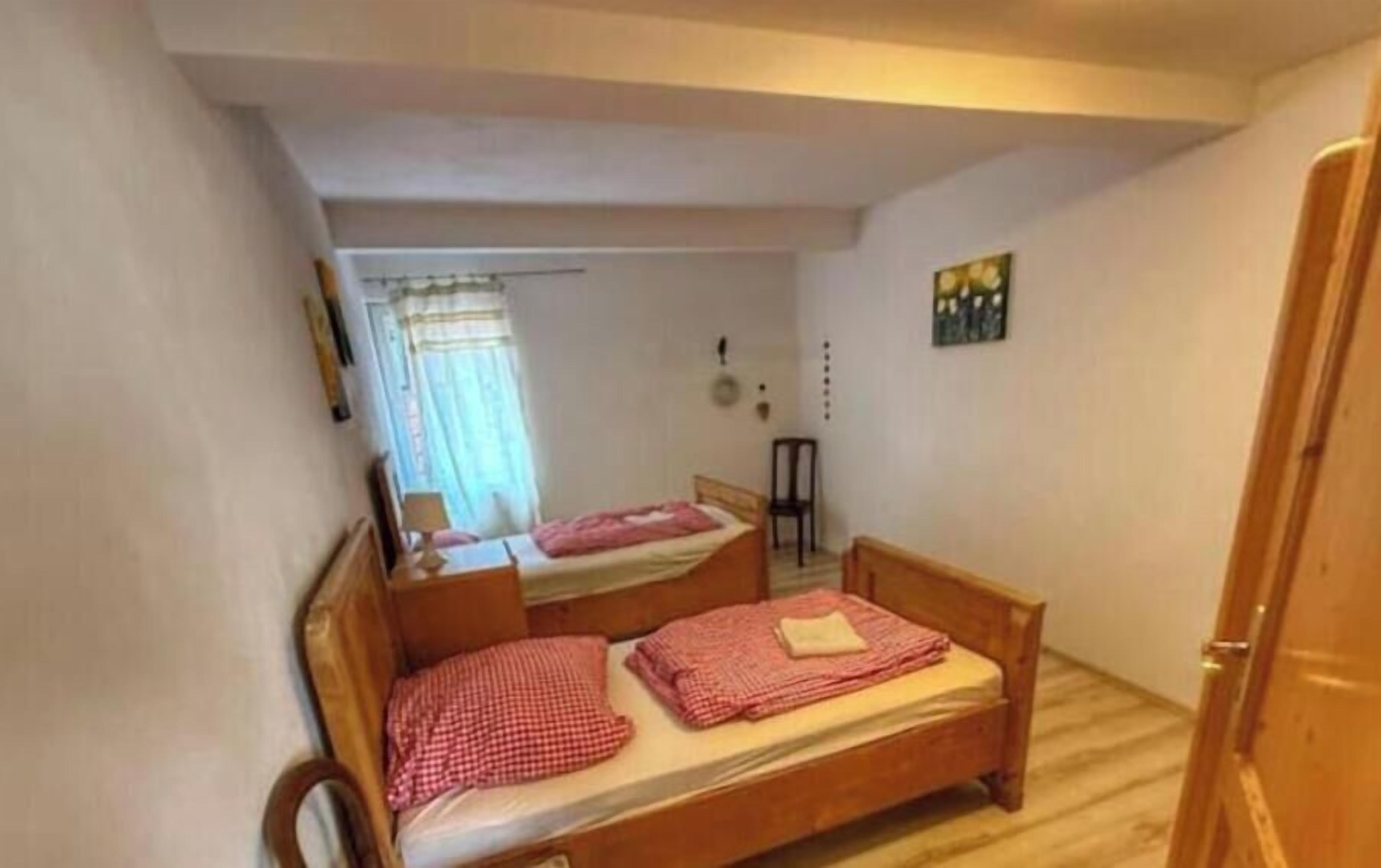 Une petite pièce avec deux lits en bois, une chaise, une porte et une fenêtre avec des rideaux.