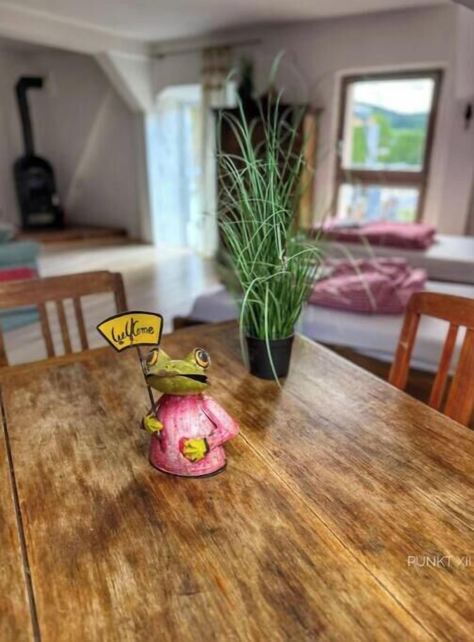 Una mesa de comedor de madera con una figurita de rana sosteniendo un letrero, una planta en maceta y una ventana al fondo.