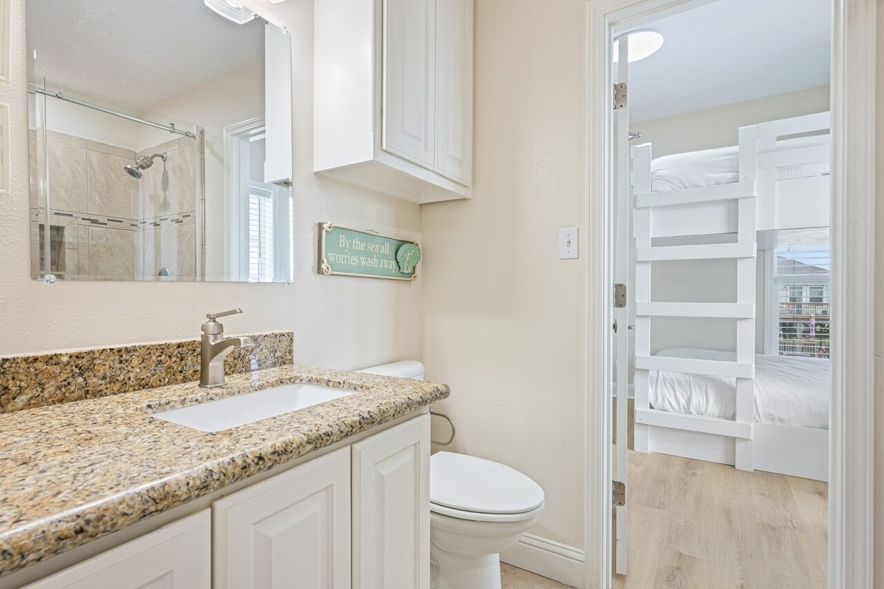 A bathroom with a granite countertop, a white sink, a shower, and a toilet. There is a sign on the wall that reads 'By the sea all worries wash away.'