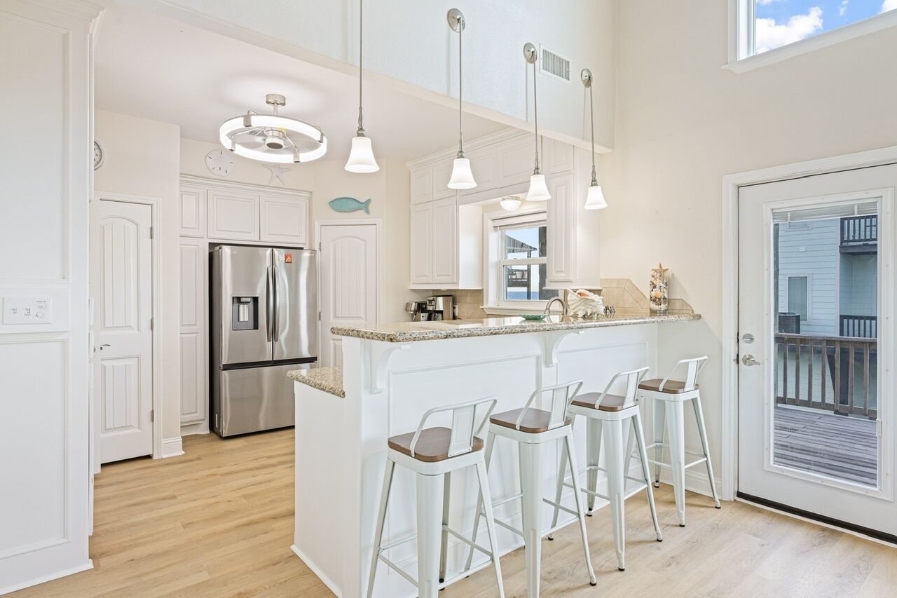 Cocina moderna con armarios blancos, refrigerador de acero inoxidable, una isla central con taburetes y luminarias colgantes, además de vistas al exterior.