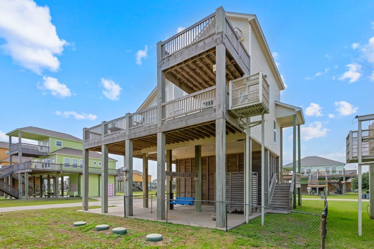 A multi-level wooden structure with a balcony, surrounded by a grassy area and other buildings.