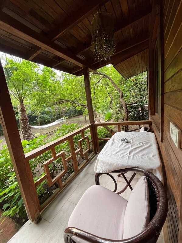 A wooden balcony with a table and chair, surrounded by lush greenery and a hammock.
