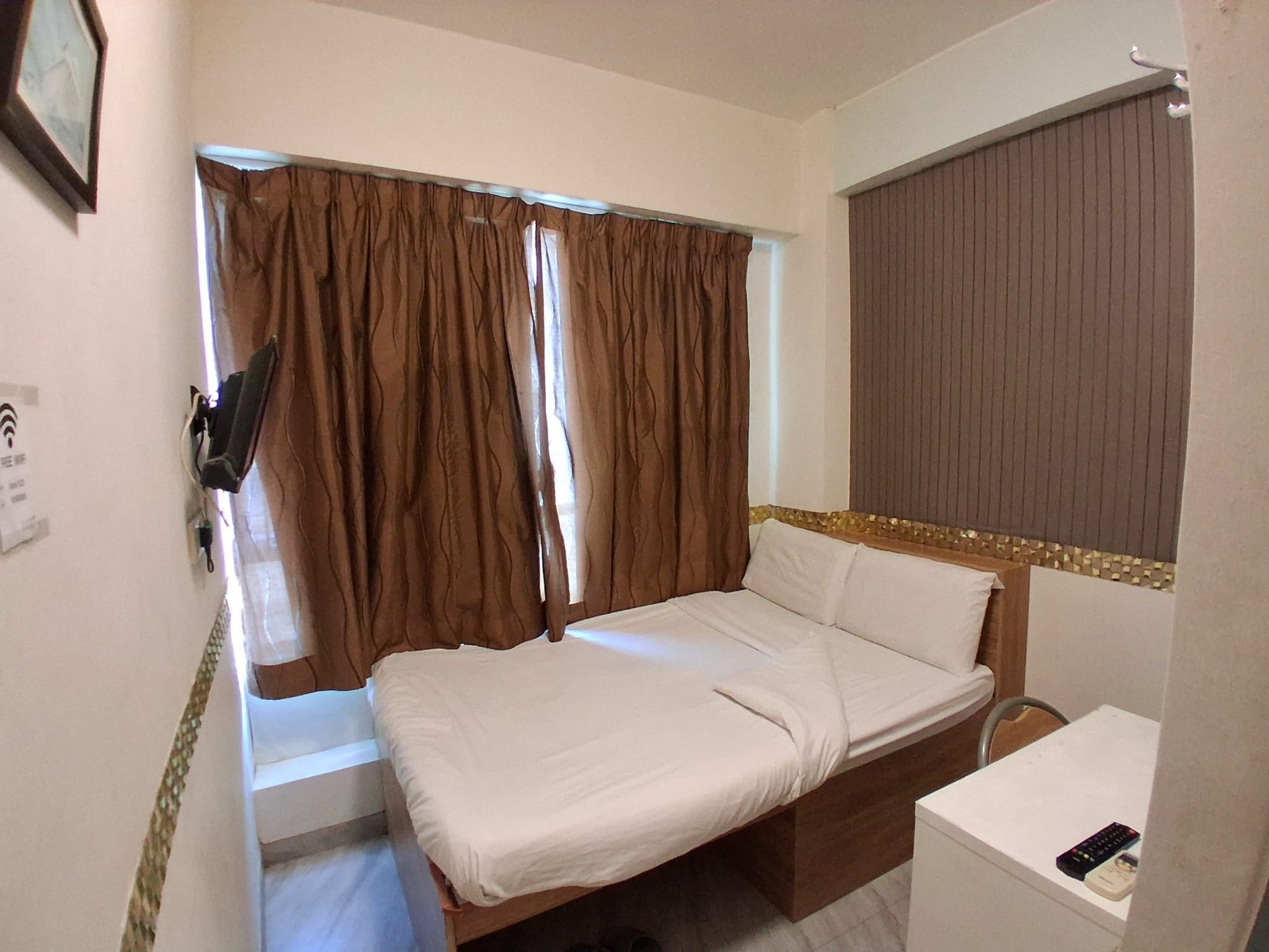 A small, single-bed room with a wooden headboard, a bedside table with a remote control, and a window with brown curtains.