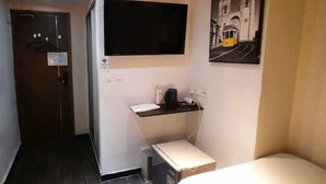 Habitación de hotel con televisor de pantalla plana montado en la pared, una mesita de noche con lámpara y un cuadro de una escena callejera.