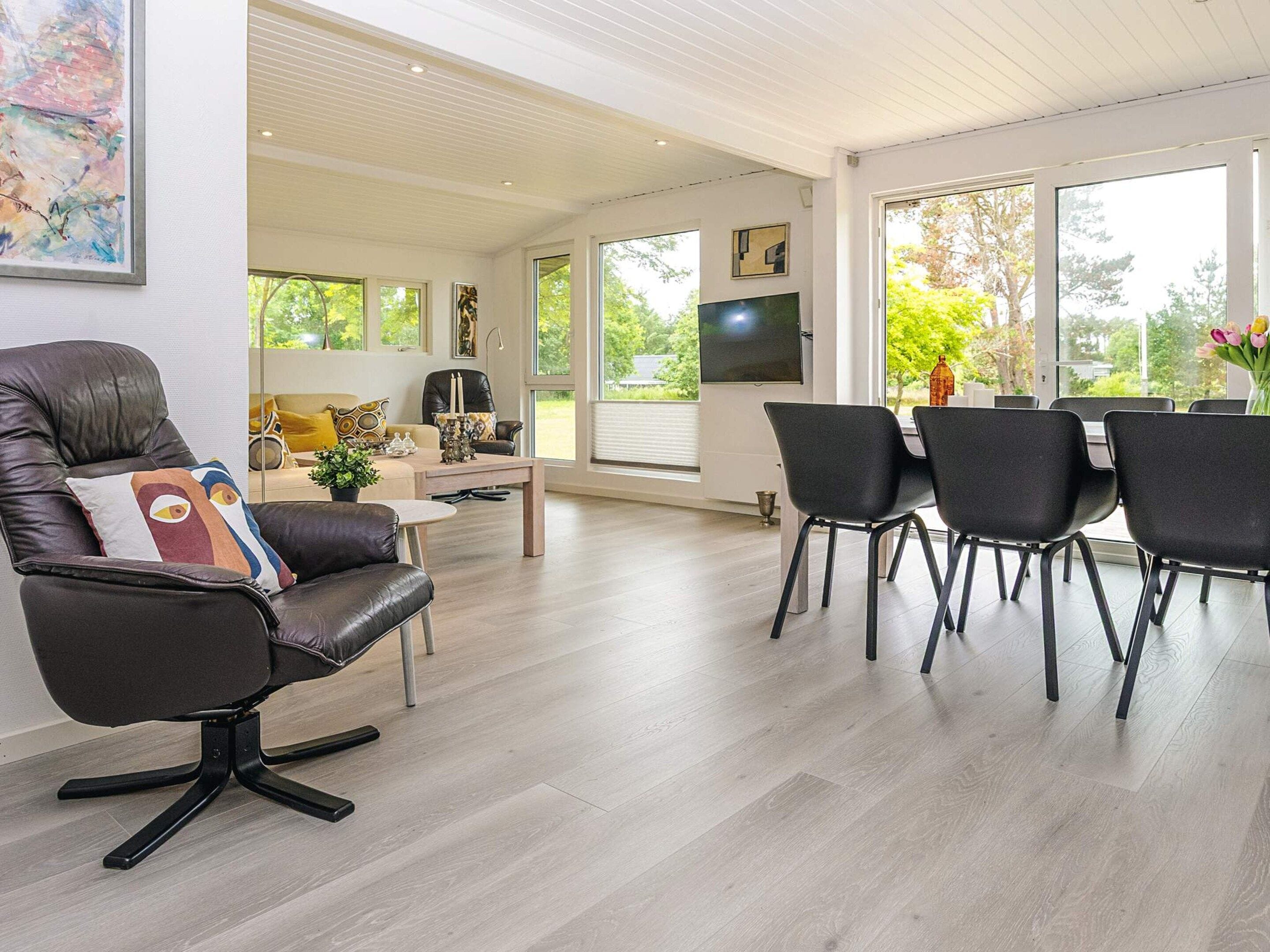 A modern living room with a leather armchair, a dining area with chairs, a flat-screen TV, and large windows offering a view of greenery.