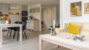 Ein modernes Wohnzimmer mit Essbereich, einer dahinterliegenden Küche und einem Sofa mit gelbem Kissen.