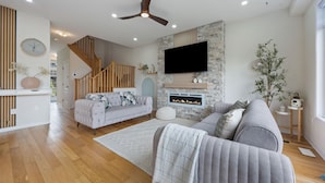 A modern living room with a fireplace, a TV mounted on the wall, and a staircase leading to an upper level. - The Triple Nest - Where Your Summer Story Begins (Hamilton)