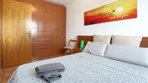 A hotel room with a bed, a wooden wardrobe, a nightstand with a green bottle, and a wall with a sunset painting. - Berni's House Palafolls Apartment with Sea Views, Private Terrace & Air Conditioning (Palafolls)