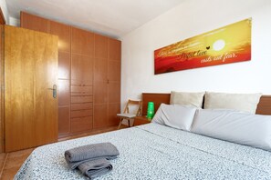A hotel room with a bed, a wooden wardrobe, a nightstand with a green bottle, and a wall with a sunset painting.