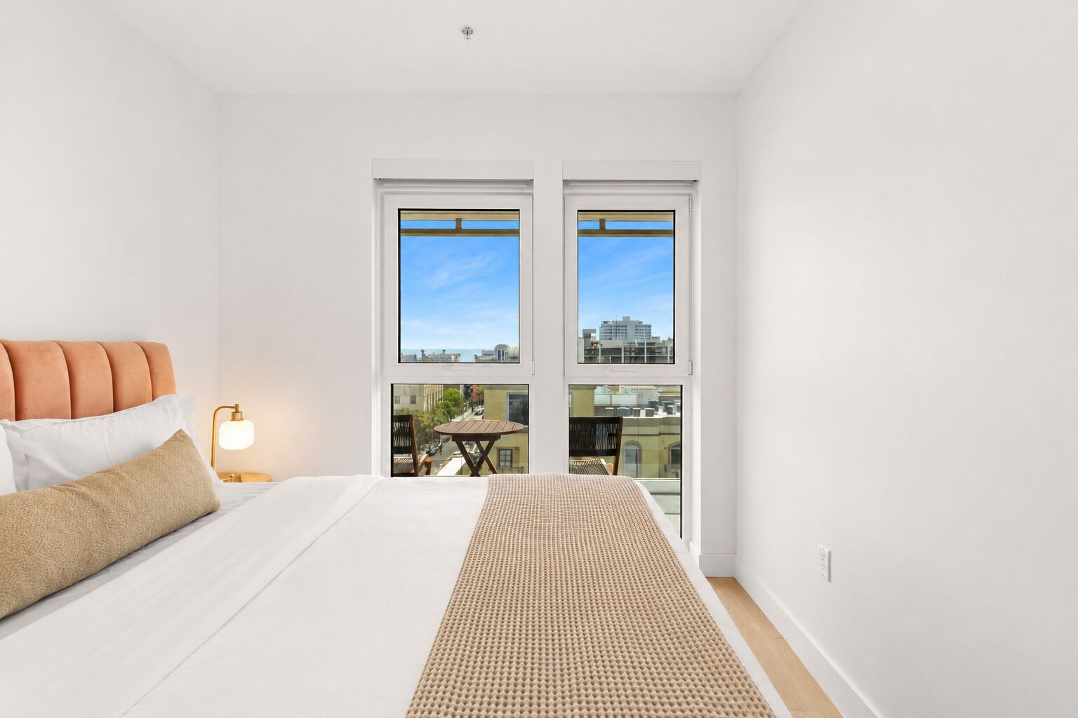 A bedroom with a bed, pillows, a bedside lamp, and a view of the city through large windows.
