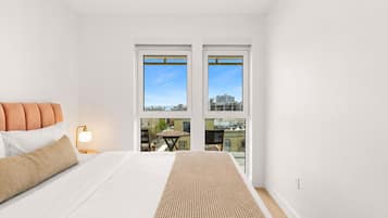 A bedroom with a bed, pillows, a bedside lamp, and a view of the city through large windows.