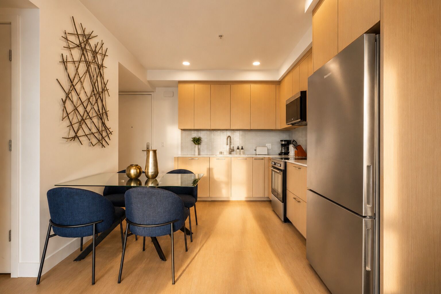 A modern kitchen with wooden cabinets, a stainless steel refrigerator, and a dining area with blue chairs.