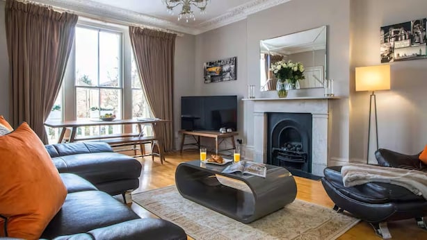 A living room with a sofa, ottoman, TV, fireplace, and a dining area with a table and chairs. - Luxury St Margarets Flat Richmond Free Parking (Twickenham)
