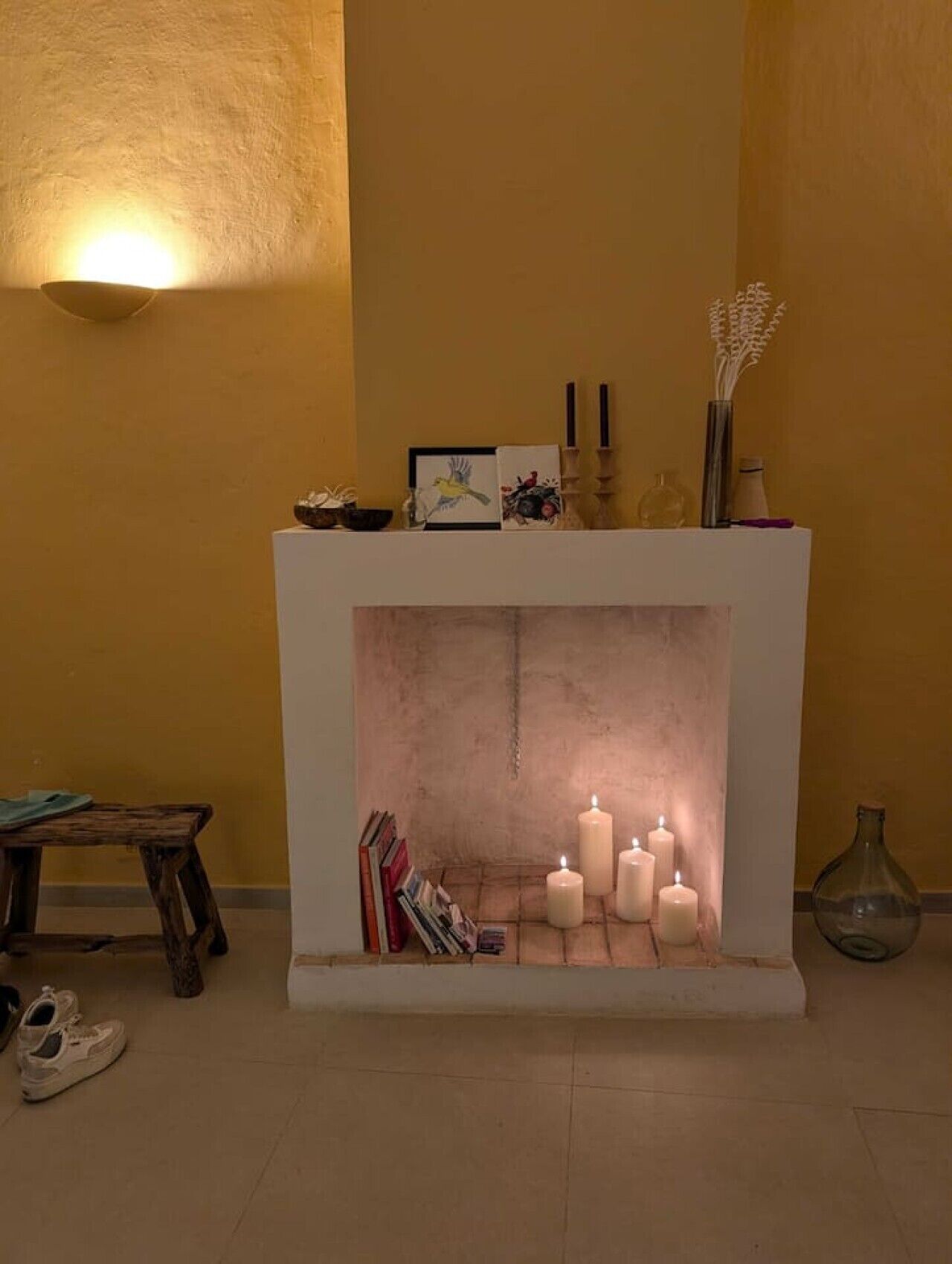 Una chimenea con velas encendidas, una mesa pequeña con libros y un jarrón en la repisa.