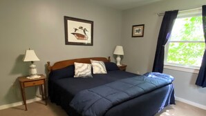 Un dormitorio con cama, dos mesitas de noche con lámparas, una ventana con cortinas y dos cuadros enmarcados en la pared.