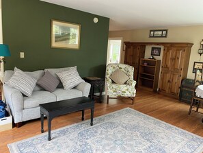 Ein Wohnzimmer mit grauem Sofa, einem gemusterten Sessel, einem schwarzen Couchtisch, einem blauen Teppich mit Muster und einer Holzkommode.
