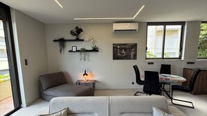 En moderne stue med en grå hjørnesofa, et lille bord med en lampe, en vægmonteret aircondition samt et spiseområde med stole og bord.