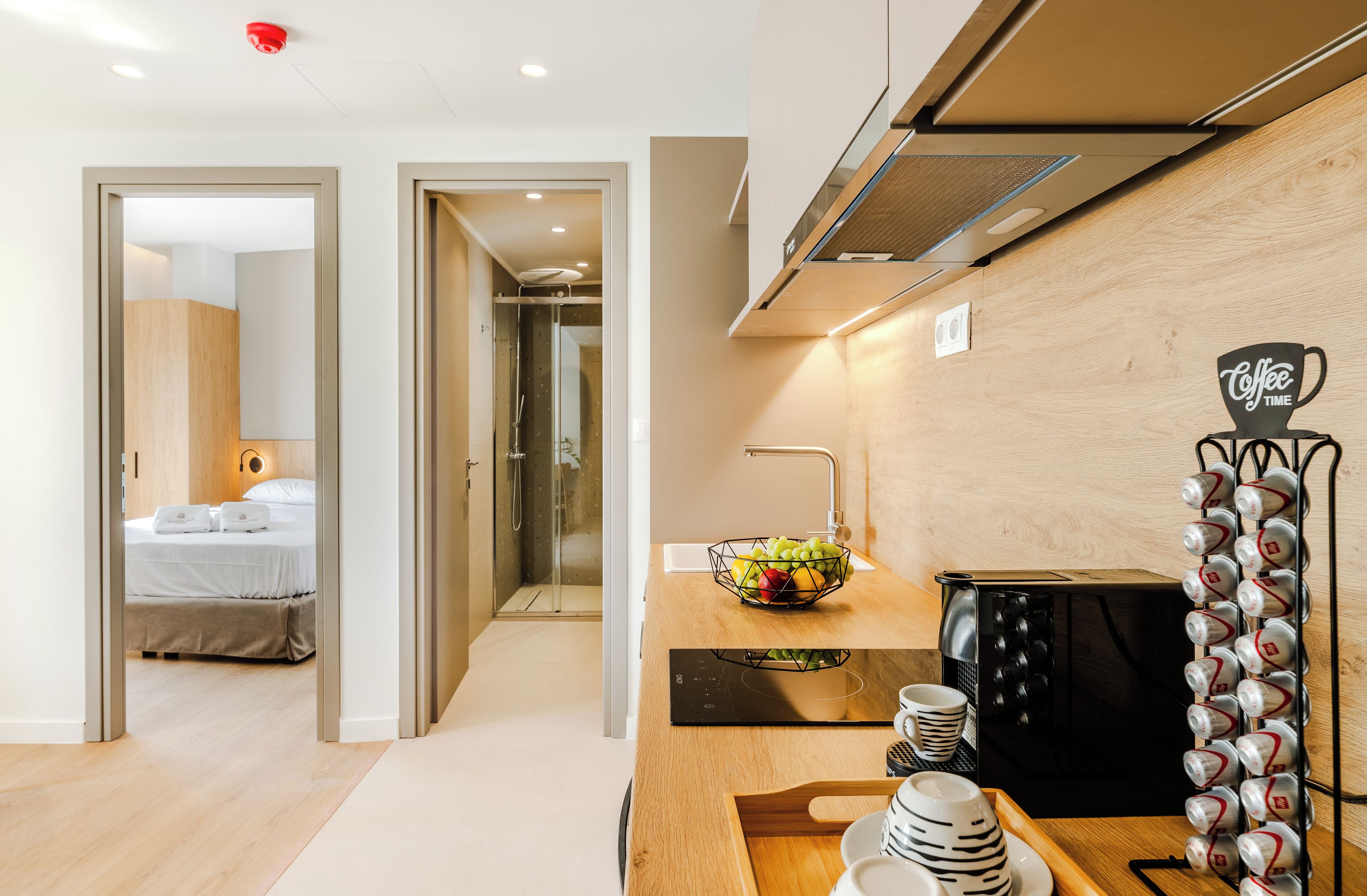 A modern hotel room with a bed, a shower, a kitchen area with a coffee machine, and a fruit bowl.