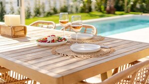 Taula de fusta per a dues persones, preparada amb plats, gots i una espelma, amb vista a una piscina.