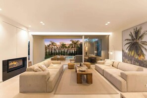 Ein modernes Wohnzimmer mit Kamin, beigen Sofas, einem Couchtisch und einem großen Fenster mit Blick auf einen Poolbereich.