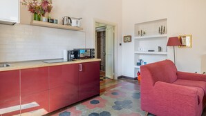 A compact kitchen with red cabinets, a microwave, and a coffee maker on the counter. There is a red sofa and a lamp in the living area. - One Bedroom Apartment (ROMA)