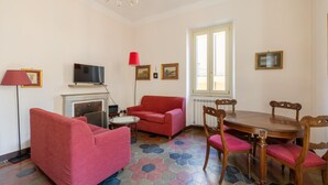 A living room with a fireplace, a television, a red sofa, a dining table with chairs, and a patterned floor. - One Bedroom Apartment (ROMA)