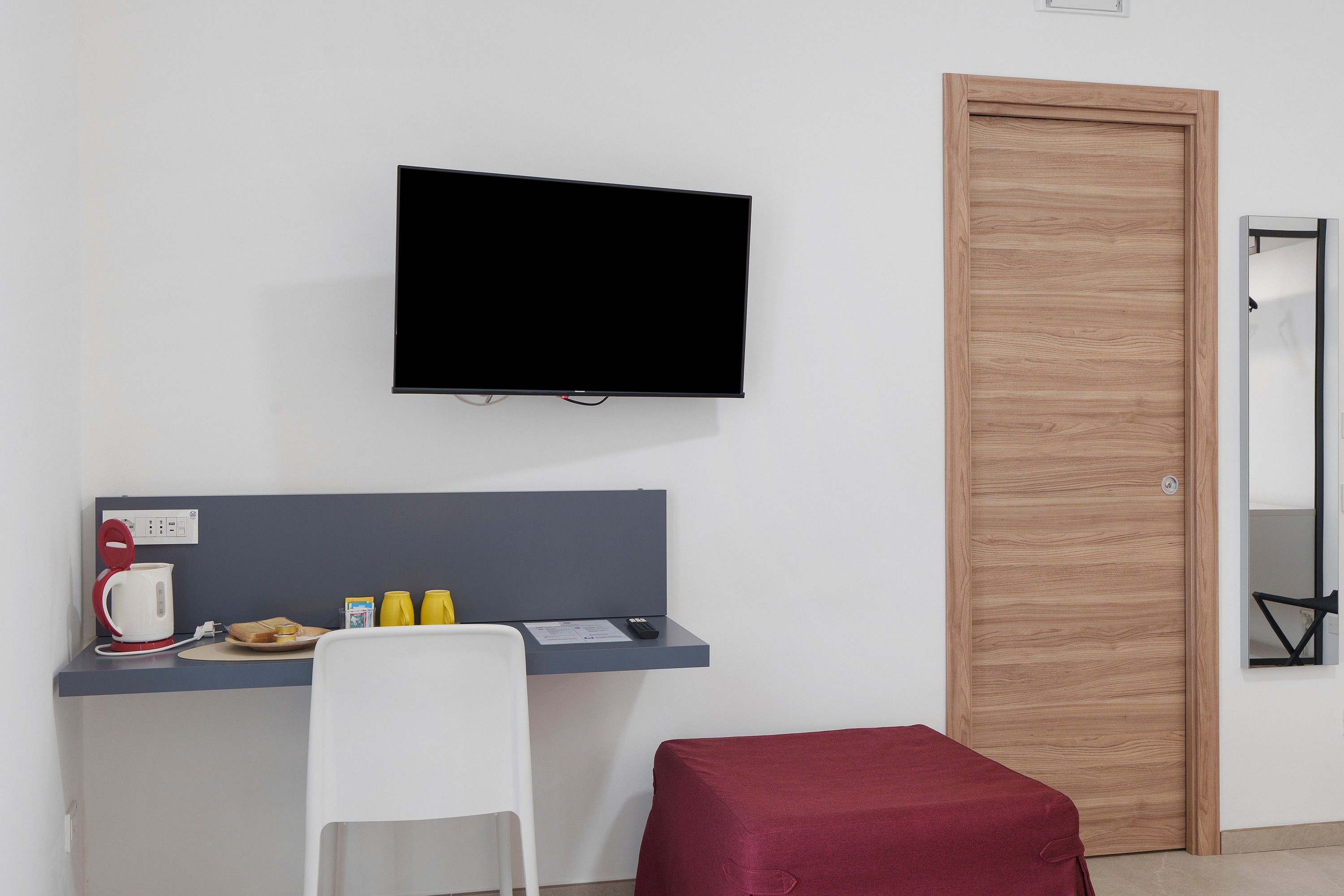 Une pièce dotée d’un téléviseur à écran plat fixé au mur, d’un bureau avec une chaise, d’un coussin rouge et d’une porte en bois.