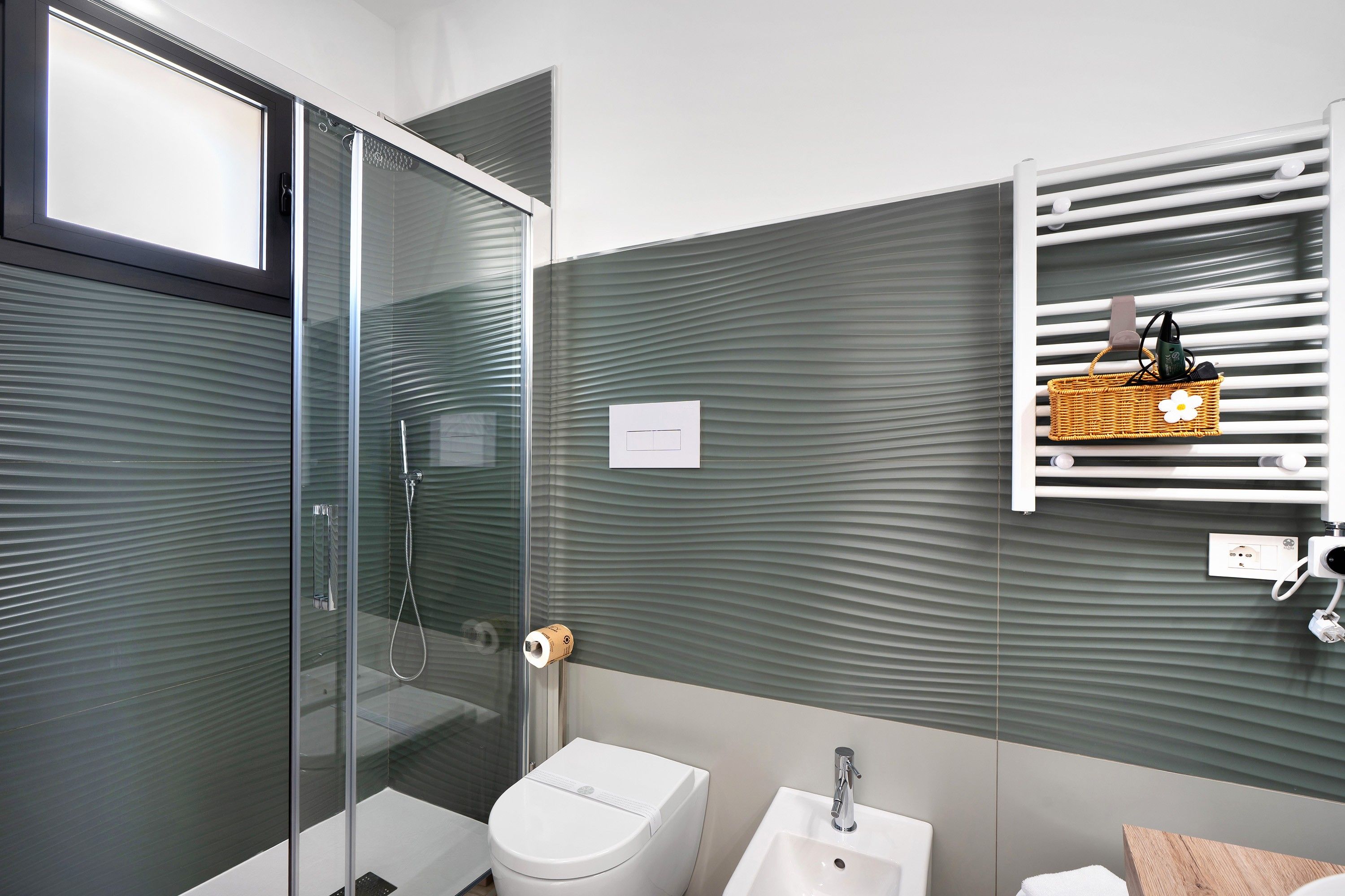 Une salle de bain moderne avec une douche à parois de verre, des toilettes blanches, un lavabo et un porte-serviettes muni d’un panier.