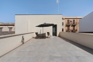 Una terraza en la azotea con mesa y sillas bajo una sombrilla, barandilla metálica y un edificio con balcones al fondo.