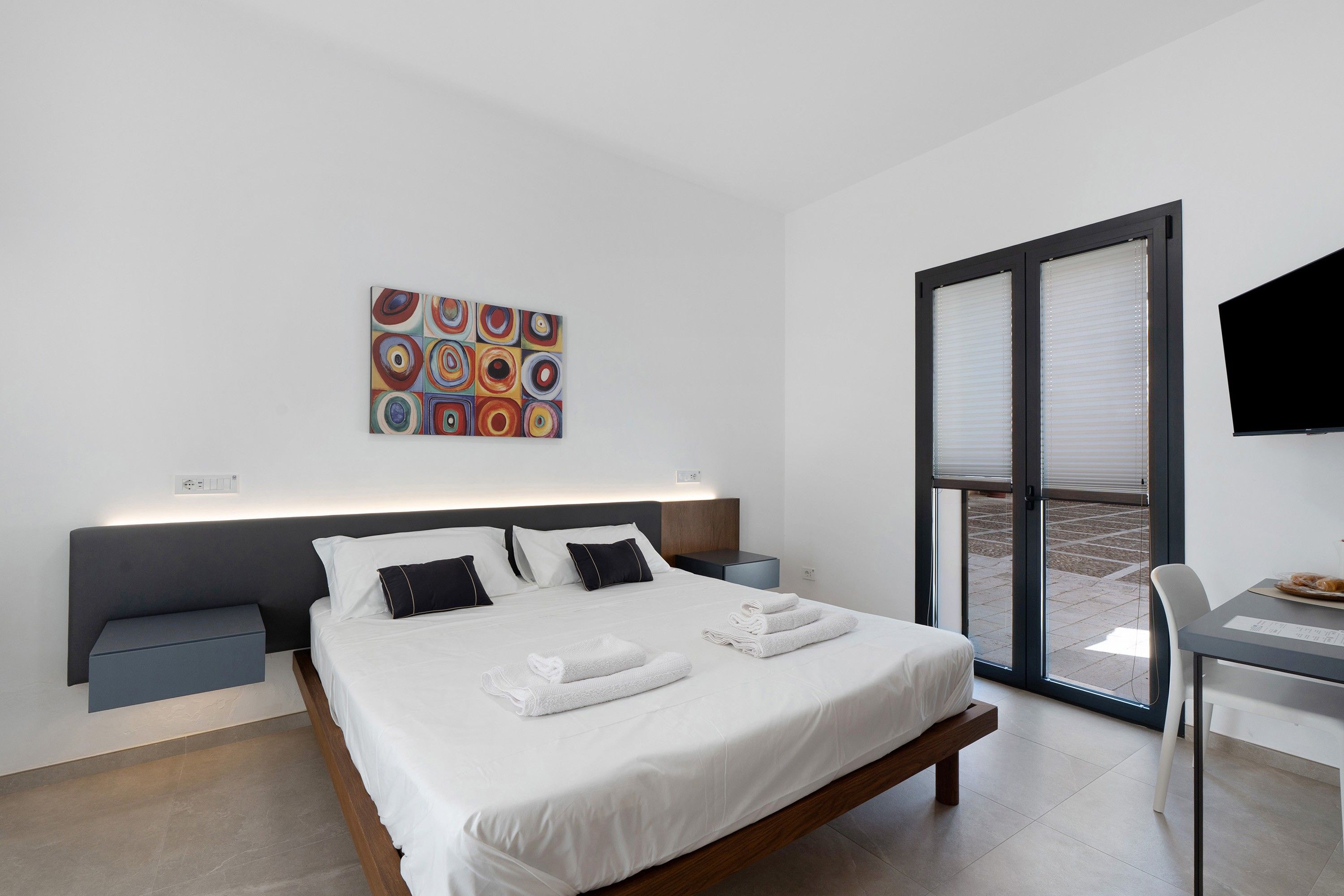 Ein modernes Hotelzimmer mit Bett, Nachttischen, Schreibtisch, Fernseher und einem großen Fenster.