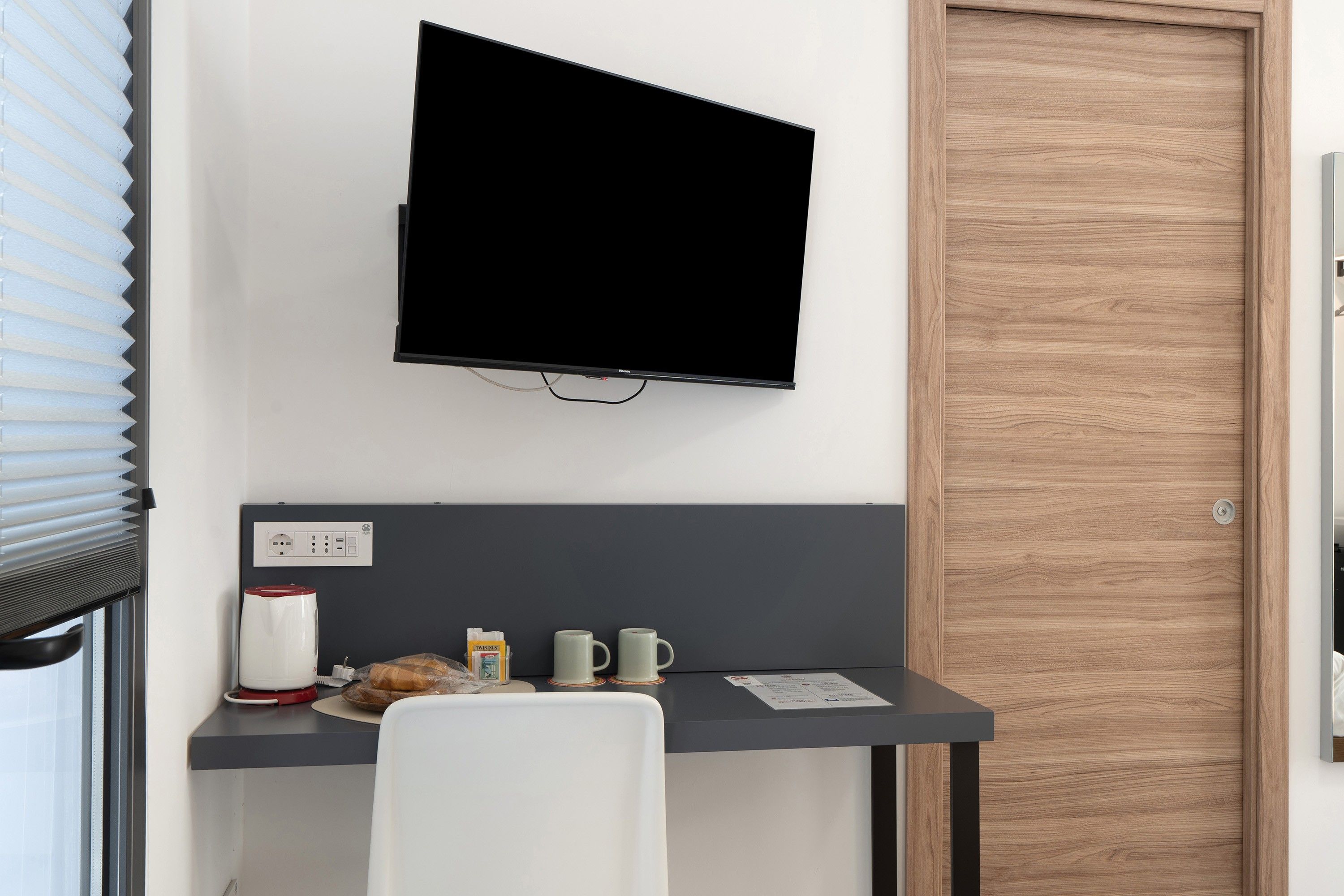 A hotel room with a flat-screen TV mounted on the wall, a desk with a coffee maker, two mugs, a plate with a pastry, and a door with a wooden panel.