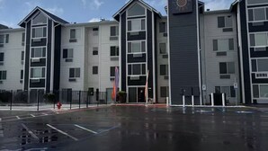 Un edificio a più piani con una torre dell'orologio centrale, un parcheggio e cielo sereno.