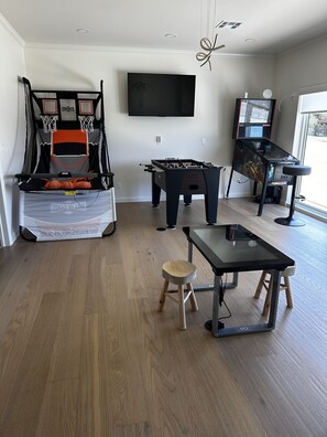 A room with a foosball table, a table tennis table, a TV mounted on the wall, and a window providing a view outside.