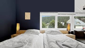 A double bed with white linens, a bedside table with a lamp, a window with a view of greenery, and a wall-mounted artwork.
