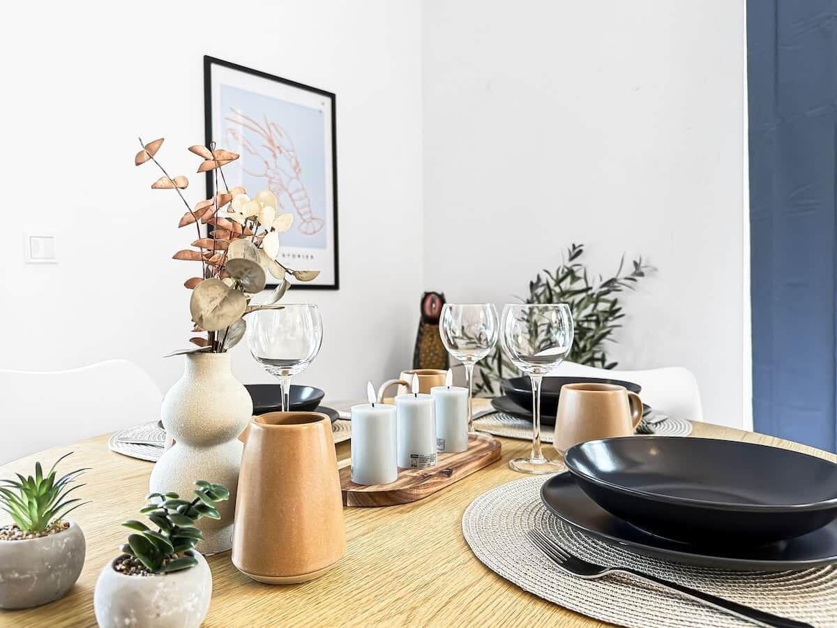 A dining table set with candles, glasses, and plates, surrounded by potted plants and a framed picture on the wall.