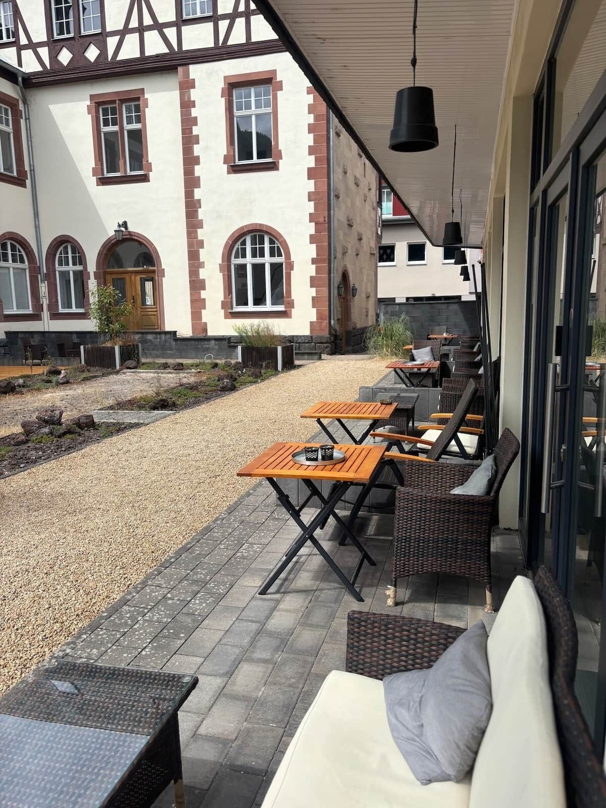 An outdoor seating area with tables and wicker chairs, a gravel path, and a building with arched windows and a wooden door.