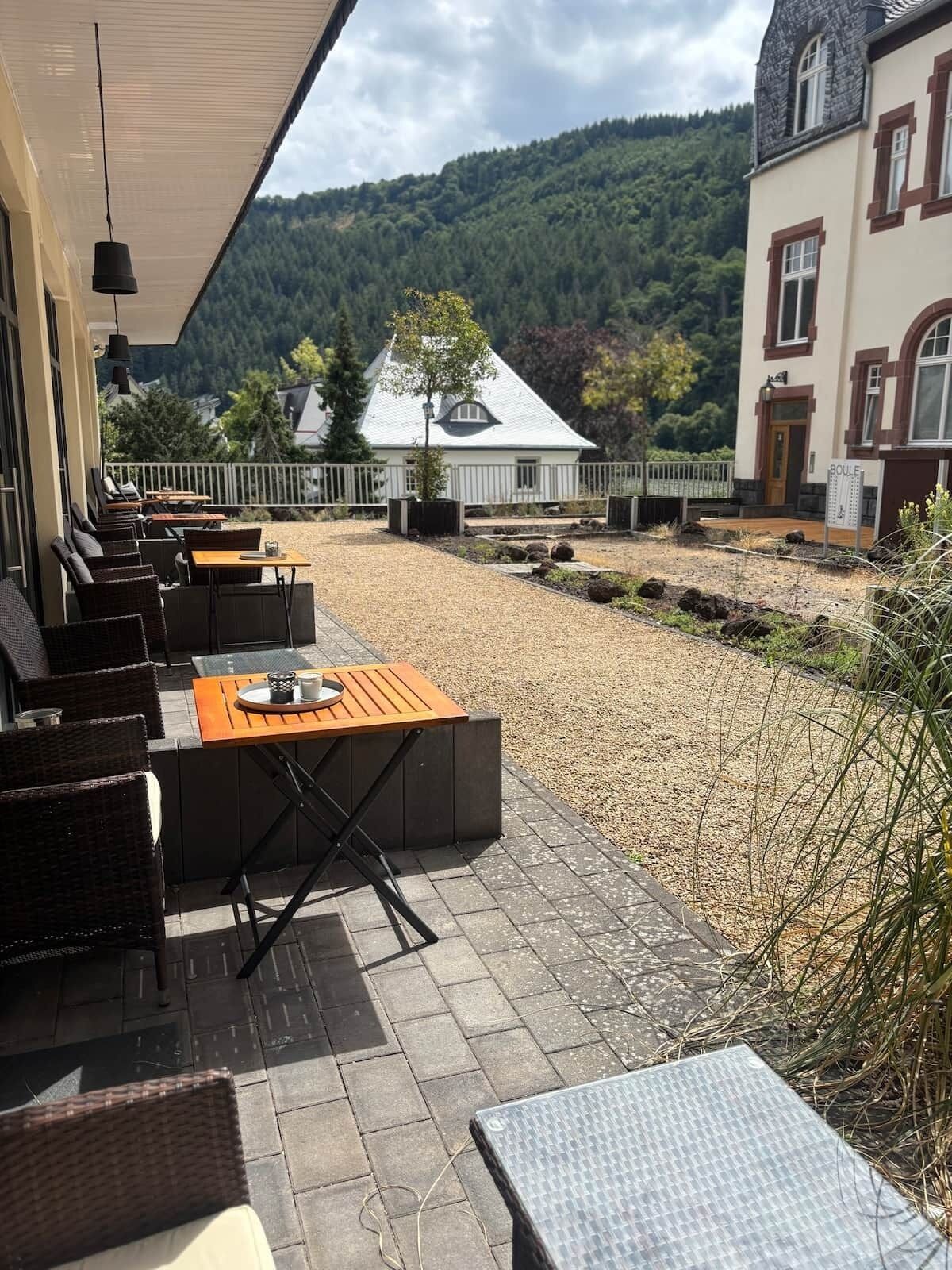 Une terrasse extérieure avec des sièges et une table, un bâtiment à la façade blanche et aux ornements rouges, et une colline boisée en arrière-plan.