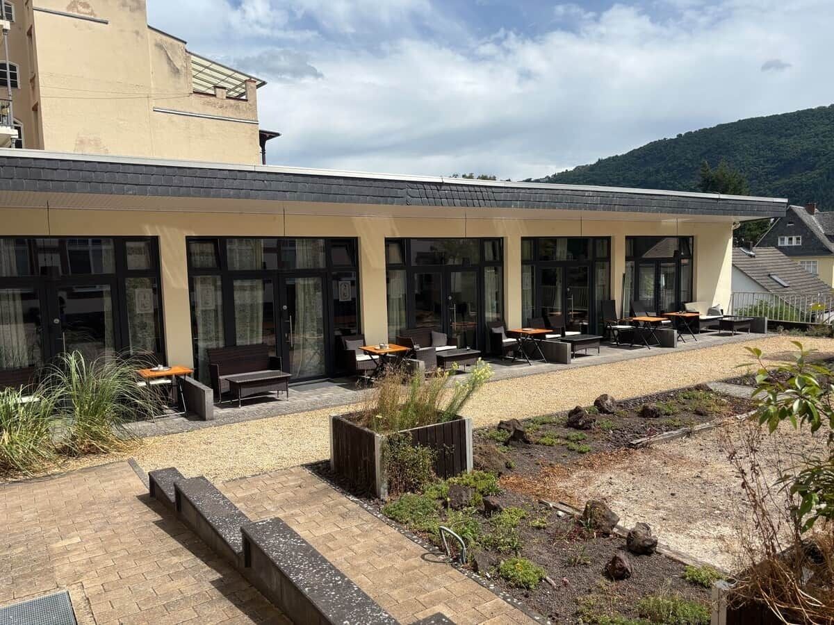 En byggnad med ett takförsett utomhusområde med sittplatser, en asfalterad gångväg och en anlagd trädgård.