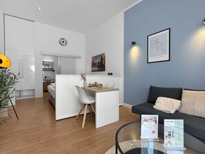 Moderni hotellihuone, jossa on pieni keittiö, ruokailutila, sohva ja lasinen pöytä, jonka päällä on esitteitä.