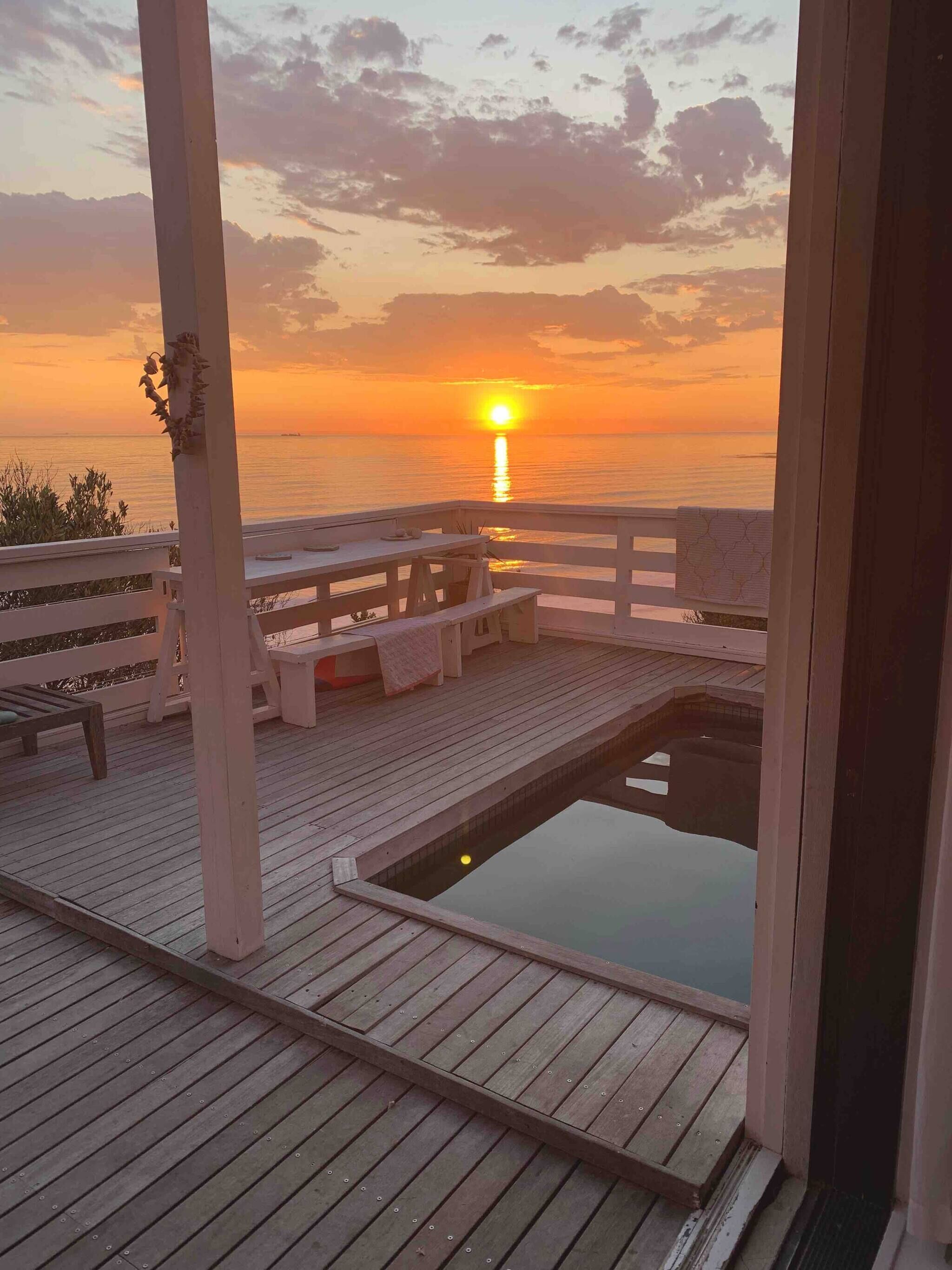 A wooden deck with a pool, benches, and a view of the sunset over the water.