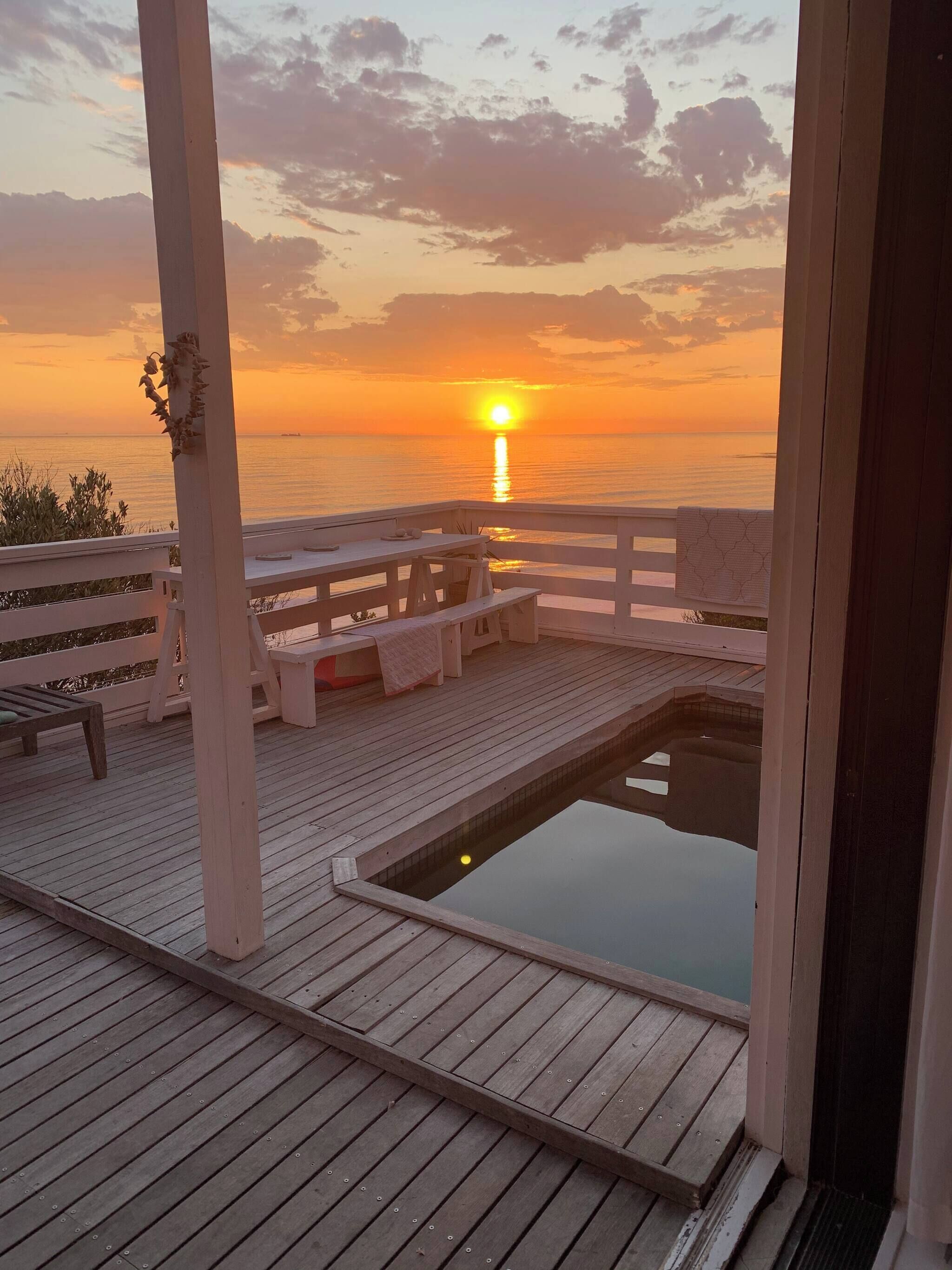 A wooden deck with a pool, benches, and a view of the sunset over the water.