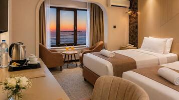 Quarto de hotel com cama, escrivaninha com cafeteira, cadeira, mesinha com bebidas e vista para o pôr do sol.