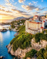 A coastal hotel with a view of the sea, surrounded by greenery and mountains in the background.
