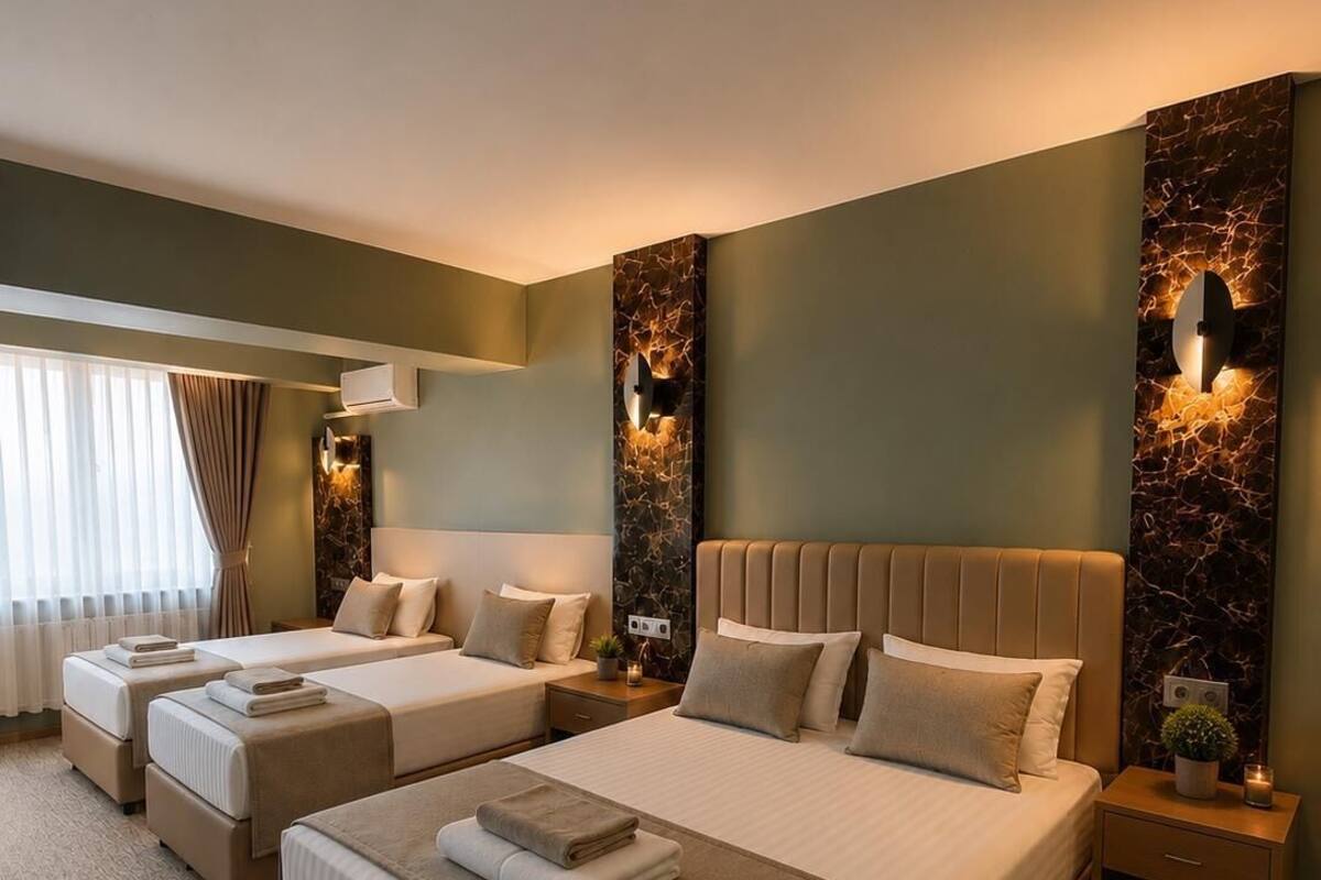 Una habitación de hotel con una cama grande, dos camas individuales, una mesita de noche con lámpara, una luz en la pared y una ventana con cortinas.