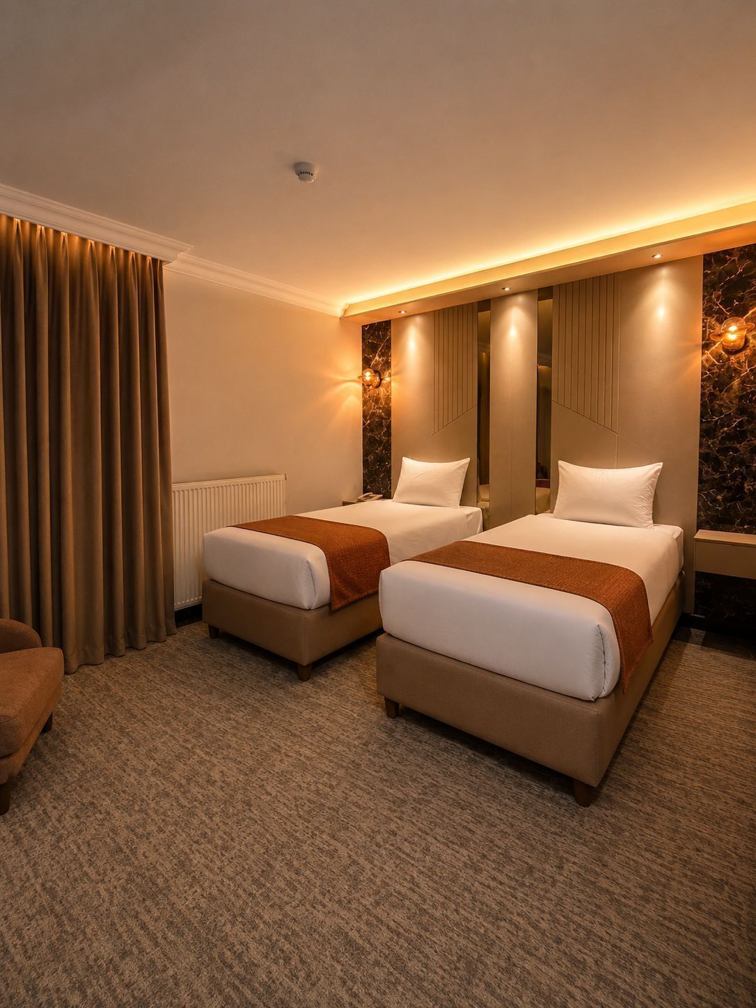 A hotel room with two beds, a radiator, a brown carpet, and a brown curtain.