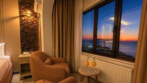 Una habitación de hotel con cama, dos sofás, una mesita con bebidas, una mesita de noche, una lámpara y vistas al atardecer sobre el océano.