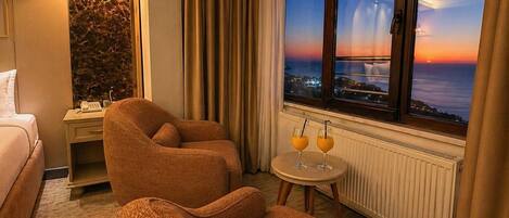 Une chambre d’hôtel avec un lit, deux canapés, une petite table avec des boissons, une table de chevet, une lampe et une vue sur le coucher de soleil sur l’océan.