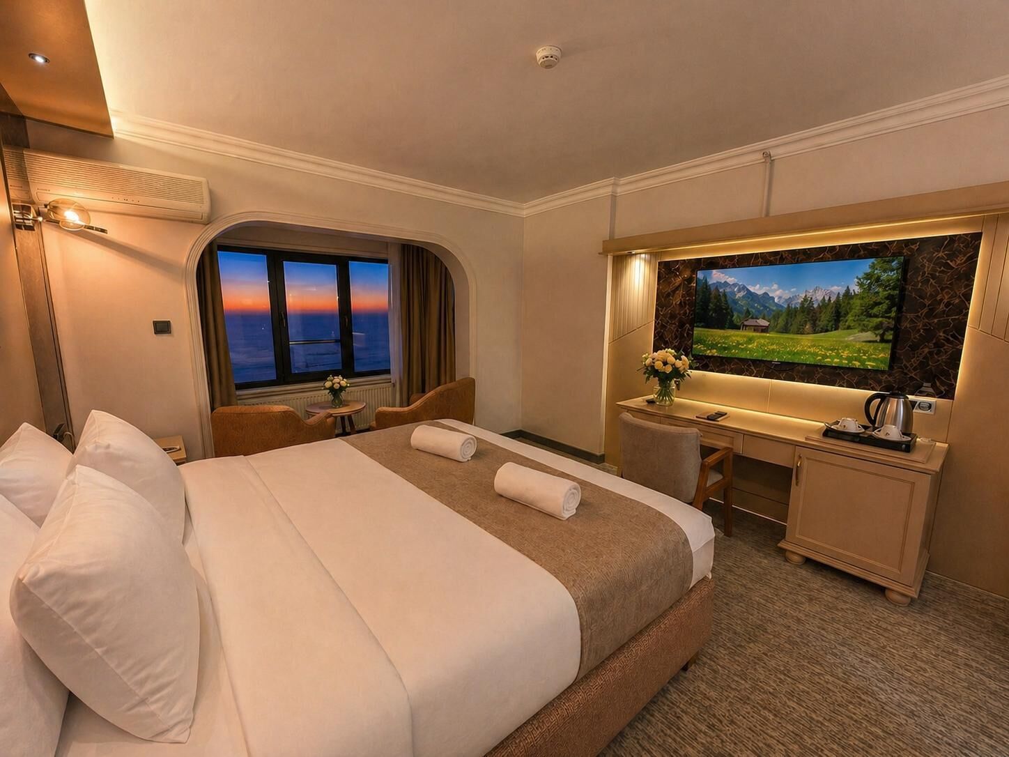 A hotel room with a bed, desk, chair, TV, and a window with a sunset view.