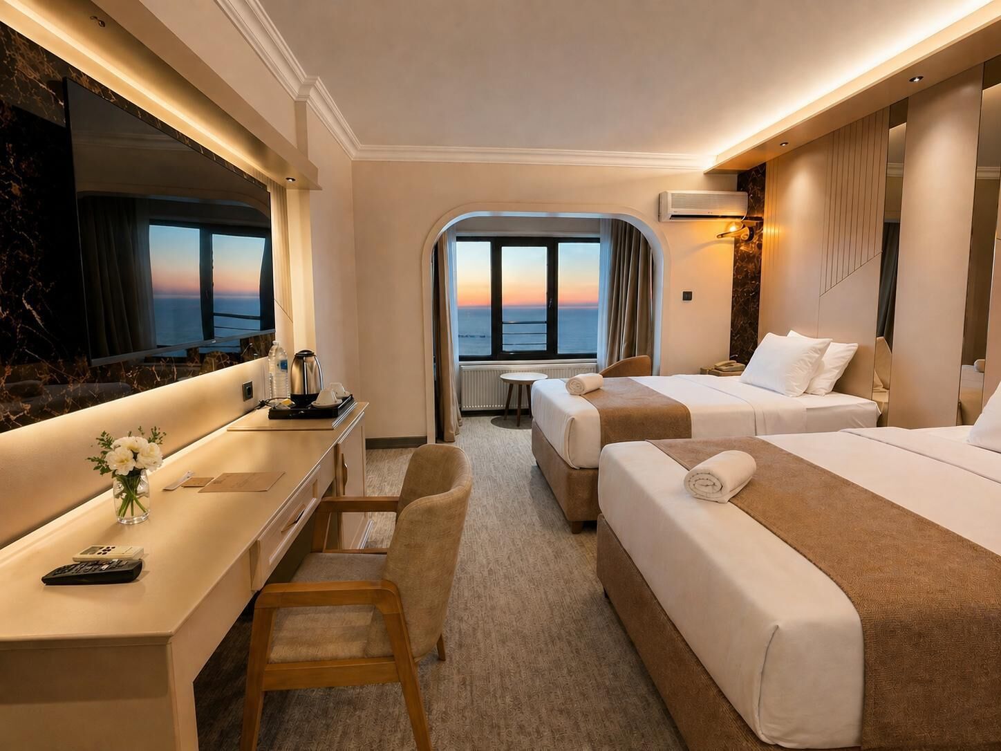 A hotel room with a desk, two beds, a view of the sea, and a large mirror.